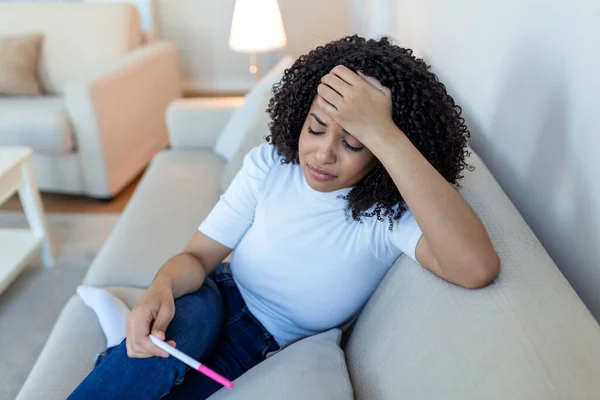 Sad, worried african american woman checking her recent pregnancy test, sitting on couch at home. Maternity, child birth and family problems concept. unwanted pregnancy