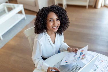 Uzaktan çalışma, teknoloji ve insan konsepti. Covid-19 sağlık krizi sırasında ev ofisinde çalışan bilgisayarlı ve evraklı mutlu gülümseyen genç iş kadını..
