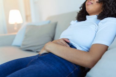 African American Woman in painful expression holding hands against belly suffering menstrual period pain, lying sad on home bed, having tummy cramp in female health concept