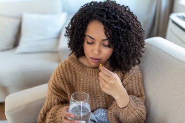 Concept of suffering from terrible pain. Beautiful, sad, upset, unhappy, troubled, weak woman with black hair, wearing casual clothes is sitting on a sofa and taking a pill