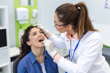 Doktor hastanın boğazını incelemek için spatula kullanıyor. KBB doktoru bir kadının boğazını muayene ediyor. Hasta boğazını kontrol etmek için ağzını açmış.