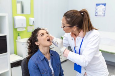 Doktor hastanın boğazını incelemek için spatula kullanıyor. KBB doktoru bir kadının boğazını muayene ediyor. Hasta boğazını kontrol etmek için ağzını açmış.