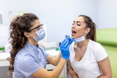 Doktor kadın hastadan boğaz temizleme testi alıyor. Sağlık çalışanı koruyucu iş elbisesinde. Salgın sırasında hastanedeler..