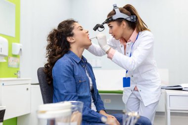 Kadın hasta, boğazına bakması için ağzını açıyor. Kadın doktor klinikteki hastanın boğazını inceliyor. Otolaryngologist hastanın boğaz ağrısını inceliyor..