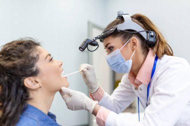 Doktor hastanın boğazını incelemek için spatula kullanıyor. KBB doktoru bir kadının boğazını muayene ediyor. Hasta boğazını kontrol etmek için ağzını açmış.