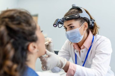 Genç bir kadın doktorunun karşısındaki muayene masasında oturuyor. Kadın kafasını kaldırıp dilini dışarı çıkarırken doktor bir dil bastırıcı ile öne uzanıyor..
