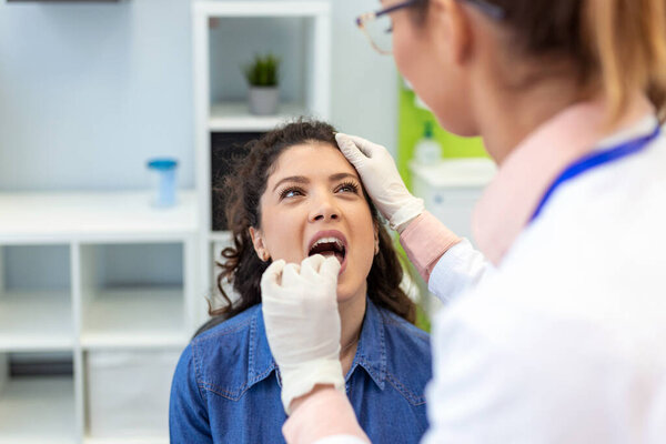 A young woman sits on an exam table across from her doctor. The doctor reaches forward with a tongue depressor as the woman looks up and sticks out her tongue.