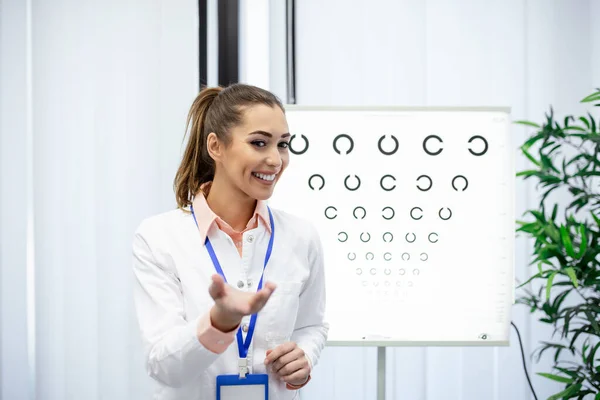 プロの女性の光学者は 目のチャートを指して ビジョンのタイムリーな診断 眼科医の肖像彼の診療所で目のチャートモニターと目の検査のために患者を求めて — ストック写真