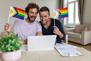 Erkek aşıkların evdeki arkadaşlarını arama videosu. Genç eşcinsel çift, video görüşmesinde arkadaşlarını selamlarken neşeyle gülümsüyor. LGBTQ bayrakları taşıyarak ve gururlu ayı birlikte kutlayarak.