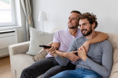 Genç homoseksüel çift oturma odasındaki kanepede dinleniyor, TV izlerken sarılıyor ve eğleniyorlar.
