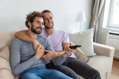 Genç homoseksüel çift oturma odasındaki kanepede dinleniyor, TV izlerken sarılıyor ve eğleniyorlar.