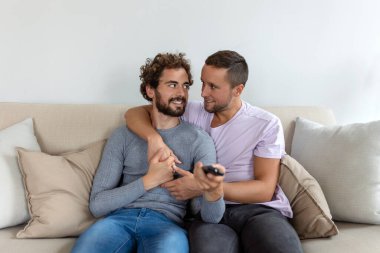 Genç homoseksüel çift oturma odasındaki kanepede dinleniyor, TV izlerken sarılıyor ve eğleniyorlar.