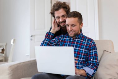 Evlerindeki oturma odalarında otururken birlikte gülen sevimli eşcinsel çift. İki romantik genç erkek aşık içeride internette sörf yaparken eğleniyor. Genç eşcinsel çift birlikte yaşıyor..