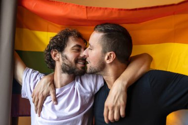 Arka planda lgbtq bayrağı taşıyan tutkulu eşcinsel çift. Genç eşcinsel çift evde sevgiyle kaynaşıyor..