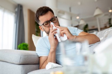 Gözlüklü hasta üzgün adam evdeki kanepede oturuyor ve burun nezlesi salgını koronavirüs salgını salgınından muzdarip. Hasta adam kötü ateş hissediyor. Virüs hastalığı belirtileri içeride.