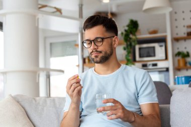 Genç adam elinde haplar ve bir bardak su tutuyordu. Sağlık ve ilaç kavramı, hasta günlük dozda reçeteli ilaç alıyor, hasta hissediyor, antibiyotik, ağrı kesici ya da antidepresan alıyor. Kapat.