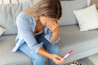 Üzgün, endişeli bir kadın son hamilelik testini kontrol ediyor, evdeki kanepede oturuyor. Doğum, doğum ve aile sorunları kavramı. istenmeyen hamilelik