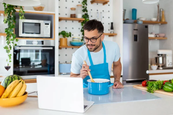 stock image Happy young man cooking healthy dinner at home. He is following a video tutorial on the laptop.