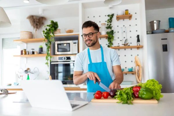 stock image Happy young man cooking healthy dinner at home. He is following a video tutorial on the laptop.