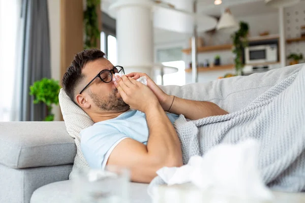 眼鏡をかけた病気の悲しい男は 自宅でソファの上に座っている鼻水インフルエンザの病気のコロナウイルスのパンデミックが流行のくしゃみに苦しんでいます 悪い熱のウイルスの病気の症状を感じるUnwell男屋内 — ストック写真