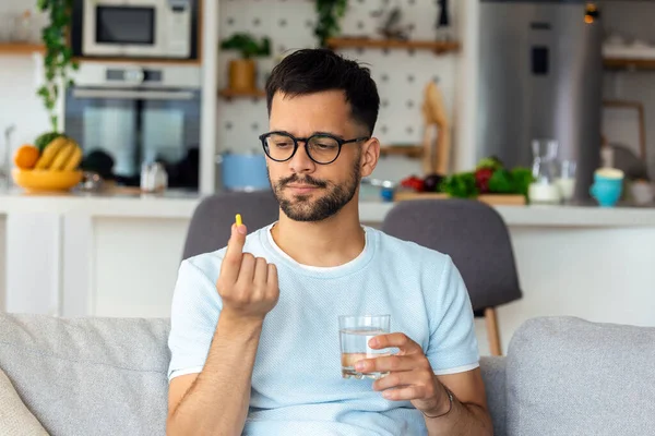 Just one pill can help. Handsome young man holding a glass of water and looking at a pill in his hand while sitting on the couch at home
