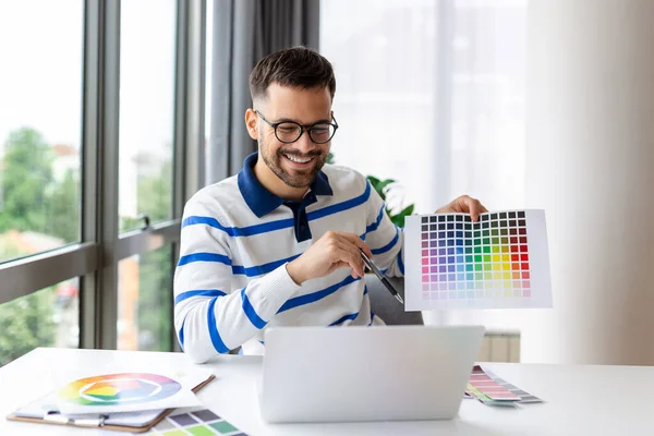 Young guy showing color swatches at laptop screen at home office, presenting graphic design project on remote meeting. Millennial designer selecting gamma for future house interior