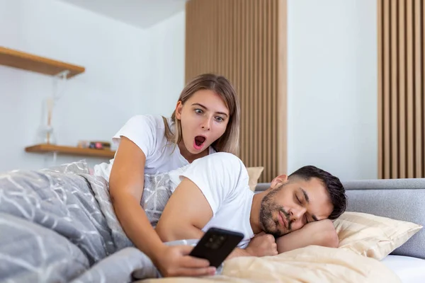 Jealous girlfriend spying the phone of her partner while he is sleeping in a bed at home. Shocked jealous wife spying the phone of her husband while he sleeping