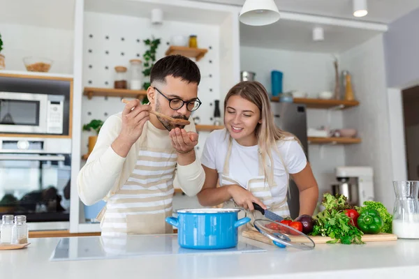 Affectionate young man kissing his wife while she's making diner. Beautiful young couple is talking and smiling while cooking healthy food in kitchen at home.