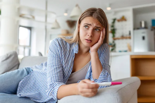 Sad, worried woman checking her recent pregnancy test, sitting on couch at home. Maternity, child birth and family problems concept. unwanted pregnancy