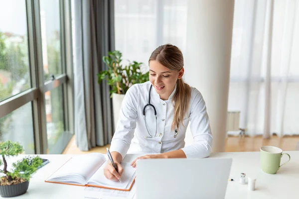 stock image Young professional female doctor wearing uniform taking notes in medical journal, filling documents, patient illness history, looking at laptop screen, student watching webinar