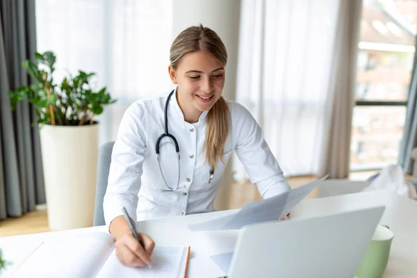 医学雑誌のノートを取り 書類を記入し 患者の病気の歴史を記入し ノートパソコンの画面を見て 学生のウェビナーを見て制服を着て笑顔のプロの女性医師 — ストック写真