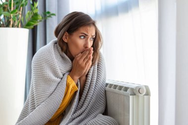 Woman freezing at home, sitting by the cold radiator. Woman with home heating problem feeling cold clipart