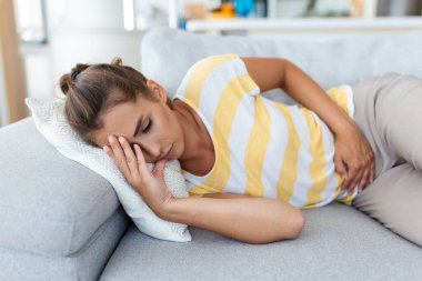 Kanepede hasta bir şekilde yatan bir kadın. Güzel genç bir kadın yatağa uzanıyor ve ellerini karnının üstüne koyuyor. Yatakta karın ağrısı çeken bir kadın. Adet dönemi.