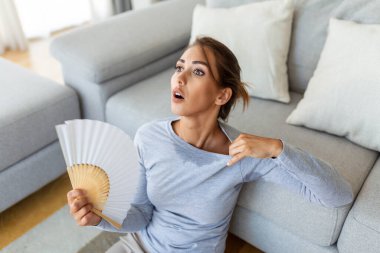 Stressed annoyed woman using waving fan suffer from overheating, summer heat health hormone problem, woman sweat feel uncomfortable hot in summer weather problem without air conditioner clipart