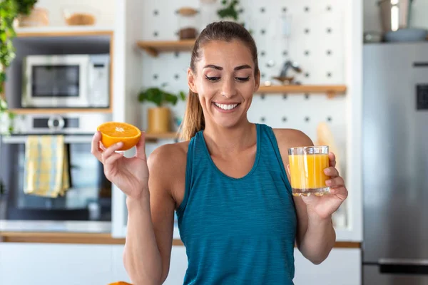 Beautiful young woman drinking fresh orange juice in kitchen. Healthy diet. Happy young woman with glass of juice and orange at table in kitchen.