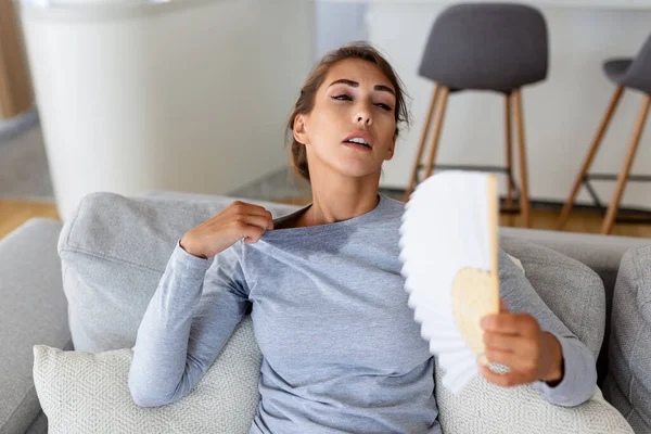 Overheated woman sit on couch at home feel warm waving with hand fan cooling down, sweating girl relax on sofa in living room hold waver suffer from heat, no air conditioner system