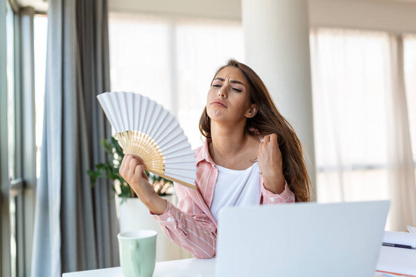 Tired overheated young woman hold wave fan suffer from heat sweating indoor work on laptop at home office, annoyed woman feel uncomfortable hot summer weather problem no air conditioner concept