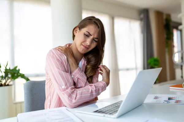 Portrait of young stressed woman sitting at home office desk in front of laptop, touching aching shoulder with pained expression, suffering from shoulder ache after working on laptop