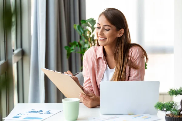Remote job, technology and people concept - happy smiling young business woman with laptop computer and papers working at home office