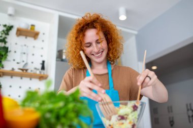 Mutfakta güzel sevimli genç gülümseyen kadın rahat giysiler içinde bir vegan salata hazırlanıyor.