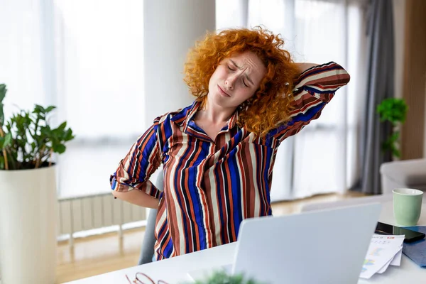 Portrait of young stressed woman sitting at home office desk in front of laptop, touching aching back with pained expression, suffering from back ache after working on laptop