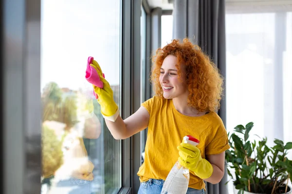 housework and housekeeping concept - happy woman in gloves cleaning window with rag and cleanser spray at home
