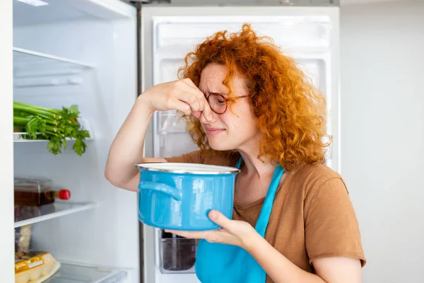 Bad Food In Fridge, young woman in holding her nose because of bad smell from food in refrigerator at home