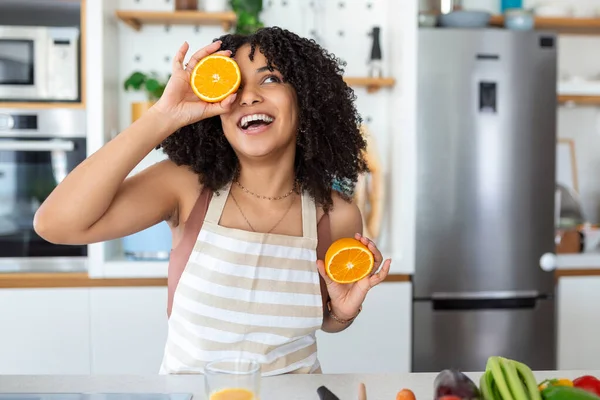 かわいいアフリカ系アメリカ人女性の笑顔と家で台所のインテリアで野菜サラダを調理しながら 2つのオレンジの部分を保持の写真 — ストック写真