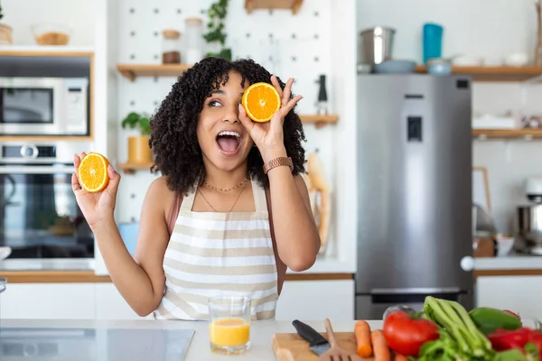 かわいいアフリカ系アメリカ人女性の笑顔と家で台所のインテリアで野菜サラダを調理しながら 2つのオレンジの部分を保持の写真 — ストック写真
