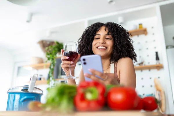Woman holding mobile phone and a glass of wine in kitchen at home