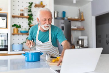 Mutlu kıdemli adam evde yemek yaparken eğleniyor. Modern mutfakta sağlıklı bir öğle yemeği hazırlayan yaşlı adam bilgisayarındaki faturaya bakıyor.