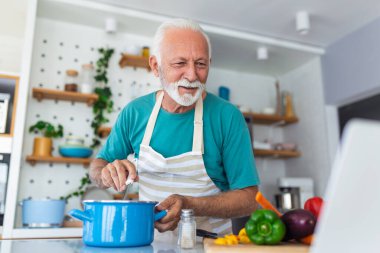Mutlu kıdemli adam evde yemek yaparken eğleniyor. Modern mutfakta sağlıklı bir öğle yemeği hazırlayan yaşlı adam bilgisayarındaki faturaya bakıyor.
