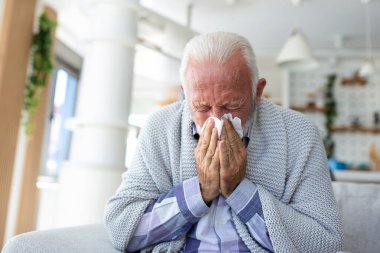 Senior sick bearded man sneezes into napkin at home on gray sofa with white blanket. Disease, protection, coronavirus, virus, disease, flu, respiratory dressing. clipart