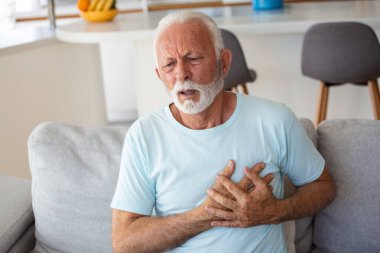 Senior man presses hand to chest has heart attack suffers from unbearable pain, Mature man with pain on heart in living room. Senior man suffering from bad pain in his chest heart attack at home clipart